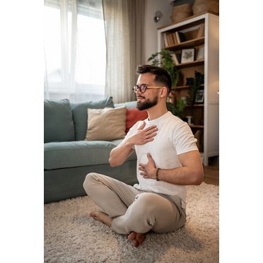 Young man practicing yoga and meditation, performing breathing exercises in lotus position on the floor in his living room, wearing casual clothes