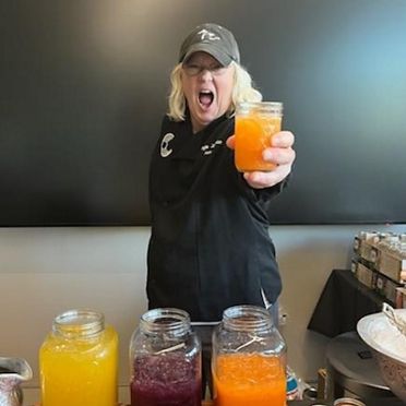 A cheerful woman in a black chef's coat and cap presents a bright orange drink with jars of colorful juices on the counter.
