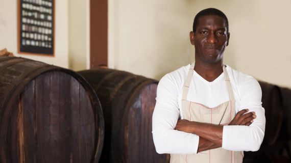 A confident black distiller in an apron stands with arms crossed in front of wooden barrels in a rustic distillery.