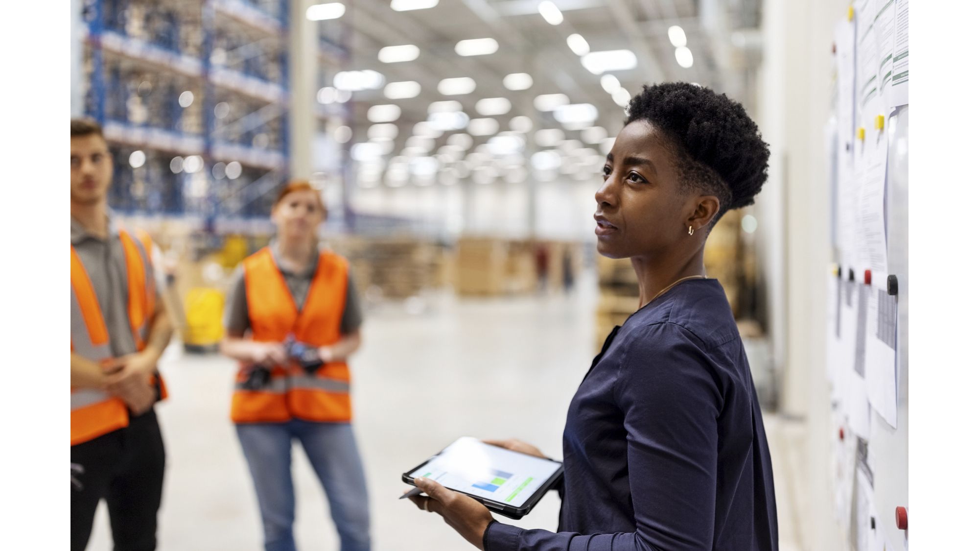 Supervisor standing by a whiteboard with a digital tablet discussing dispatch plan with workers.  Team of workers having meeting in a distribution warehouse.