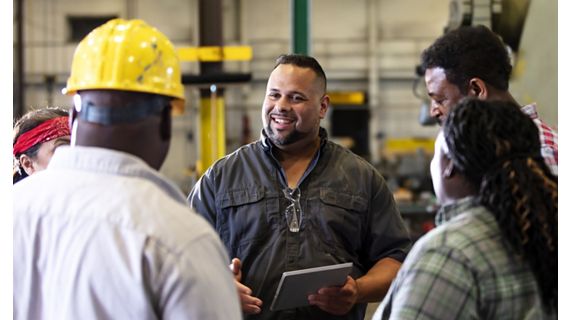 A group of five multiracial mature adults, two women and three men, working in a metal fabrication shop having a meeting on the factory floor. A mixed race Hispanic and Caucasian man is in charge, holding a digital tablet, talking and looking toward an African-American man wearing a hard hat, smiling.