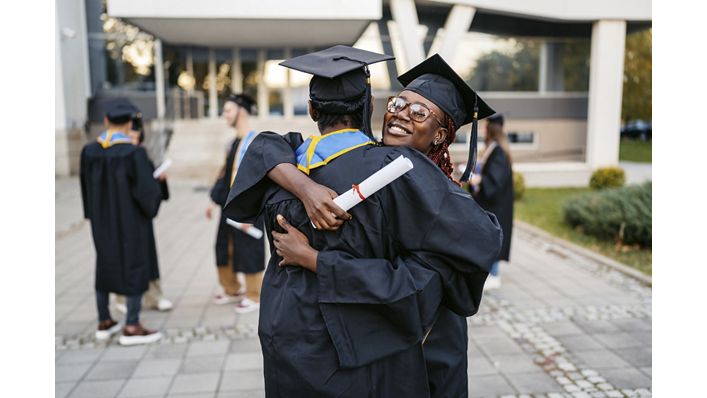 Male and female student in graduation gowns embracing each other on the university campus outdoors.