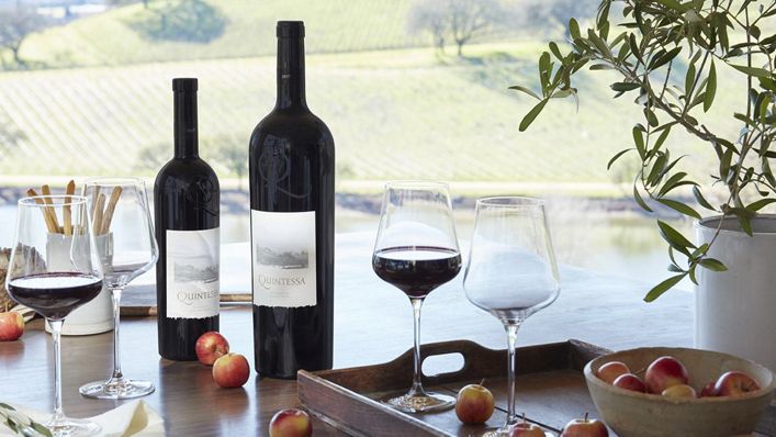 Two Quintessa wine bottles and filled glasses, amidst a tray of cheese and apples in a vineyard setting.