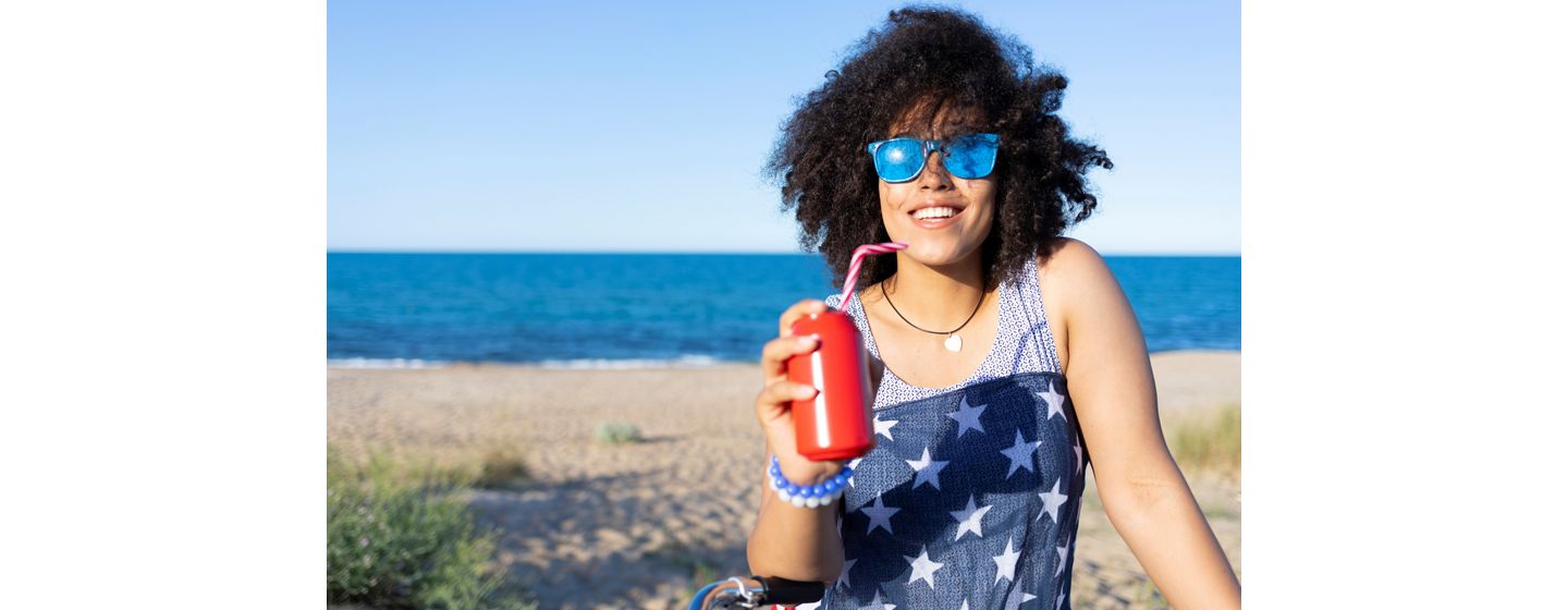 A happy woman in a star-patterned dress sipping  from a red can with a straw on a sunny beach wearing blue mirrored sunglasses.