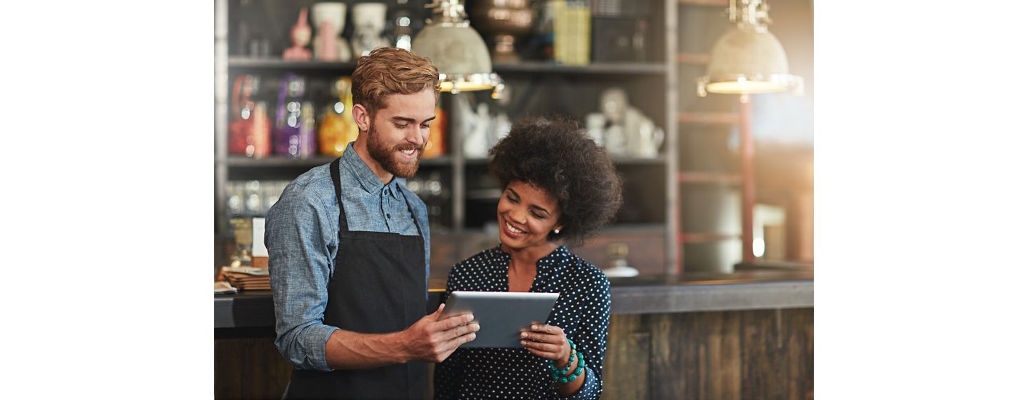 A cheerful bartender in a denim shirt and apron shares a tablet screen with a smiling sales woman with a curly afro.