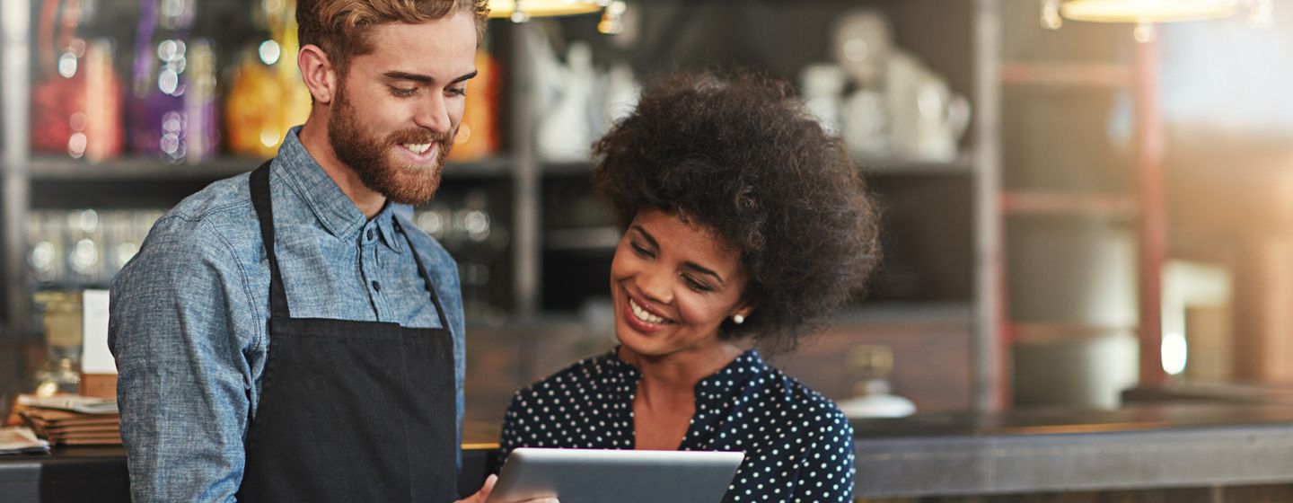 A cheerful bartender in a denim shirt and apron shares a tablet screen with a smiling sales woman with a curly afro.