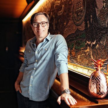 A casual portrait of mixologist Derek Brown leaning on a bar with a intricately designed mural in the background.