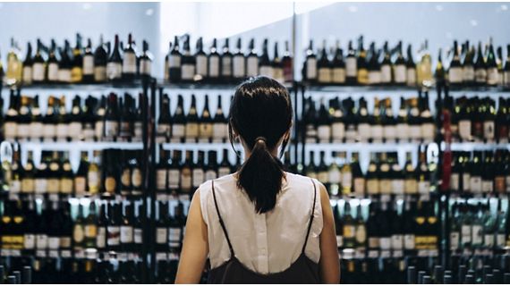 Rear view of young Asian woman grocery shopping for wines in a supermarket. She is standing in front of the liquor aisle and have no idea which wine to choose from