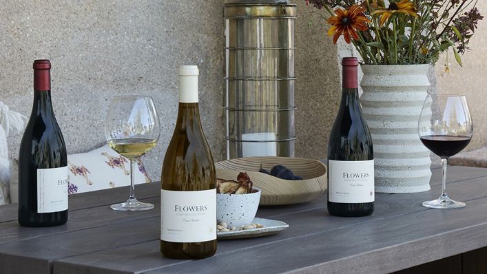An outdoor table setting featuring three wine bottles, two reds and one white, from Flowers Vineyard, accompanied by glasses of wine and a snack bowl, with a warm and inviting ambiance.