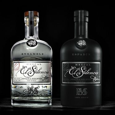 Two bottles of El Silencio Mezcal, one clear and one dark, stand out against a black background.