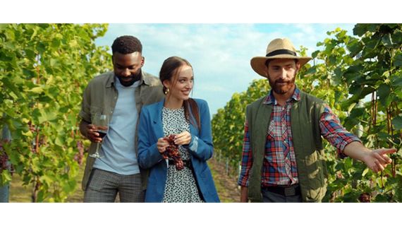 Group of people in a vineyard with one holding a glass of wine and a woman holding a cluster of grapes