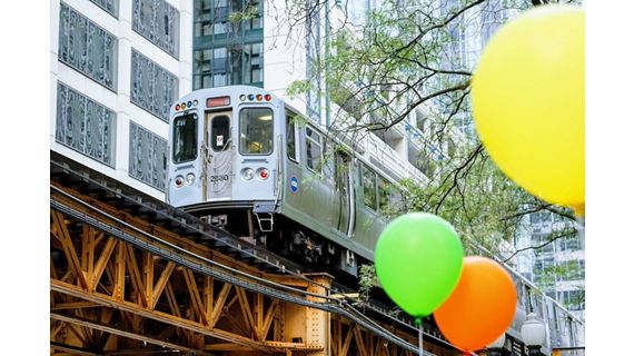 An urban scene with a focus on a Chicago El train moving along the elevated tracks, with vibrant green and orange balloons in the foreground.