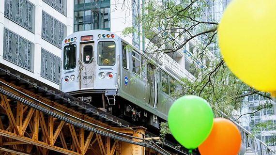An urban scene with a focus on a Chicago El train moving along the elevated tracks, with vibrant green and orange balloons in the foreground.