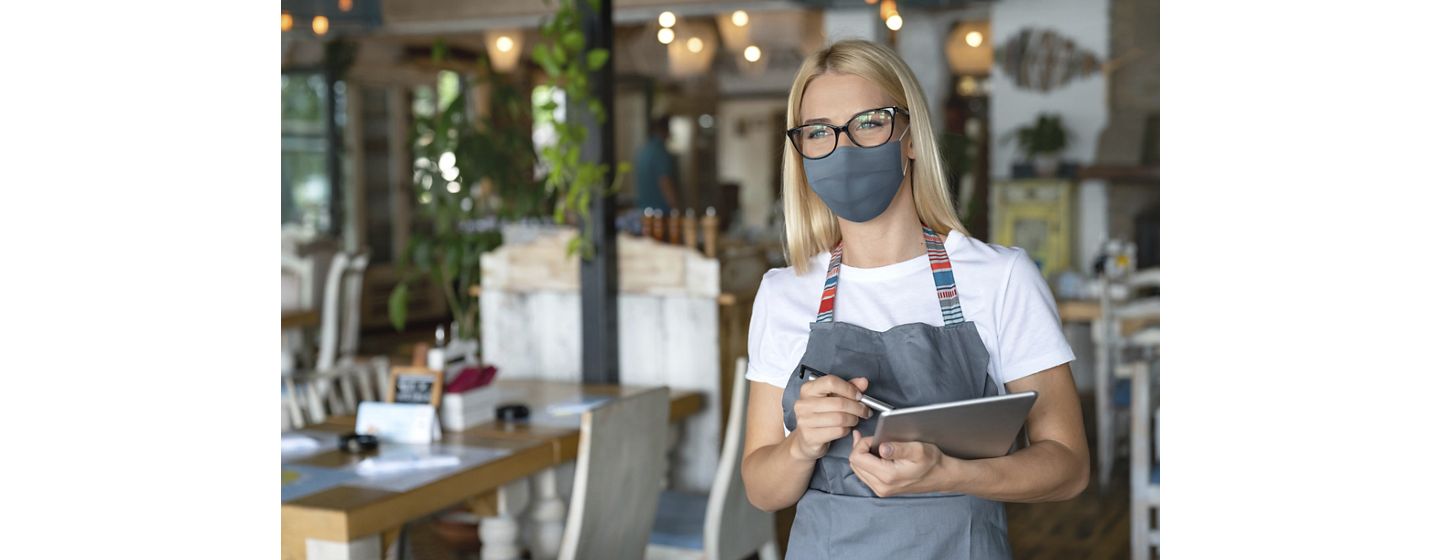 Female waiter wearing an apron, glasses and face mask with a pen and tablet in her hand