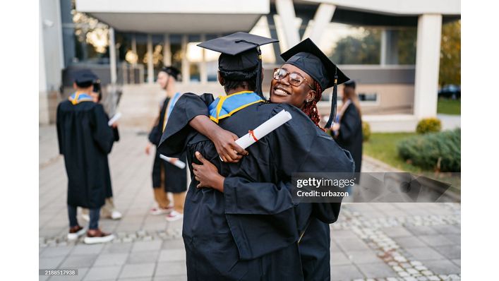 Male and female student in graduation gowns embracing each other on the university campus outdoors.