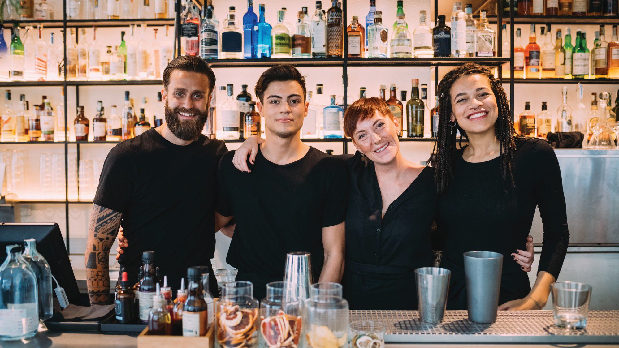 A happy team of bartenders standing behind a bar, smiling confidently at the camera.