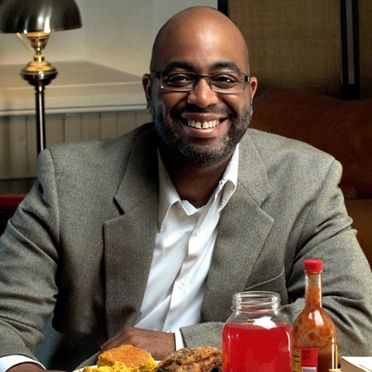 A smiling man in glasses with a short beard, wearing a smart grey suit, seated in a restaurant.