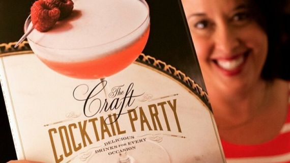 A smiling woman holding a cocktail-themed book titled The Craft Cocktail Party: Delicious Drinks for Every Occassion.