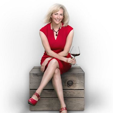 A portrait of Natalie Maclean seated on a wooden crate, wearing a red dress, holding a glass of red wine.