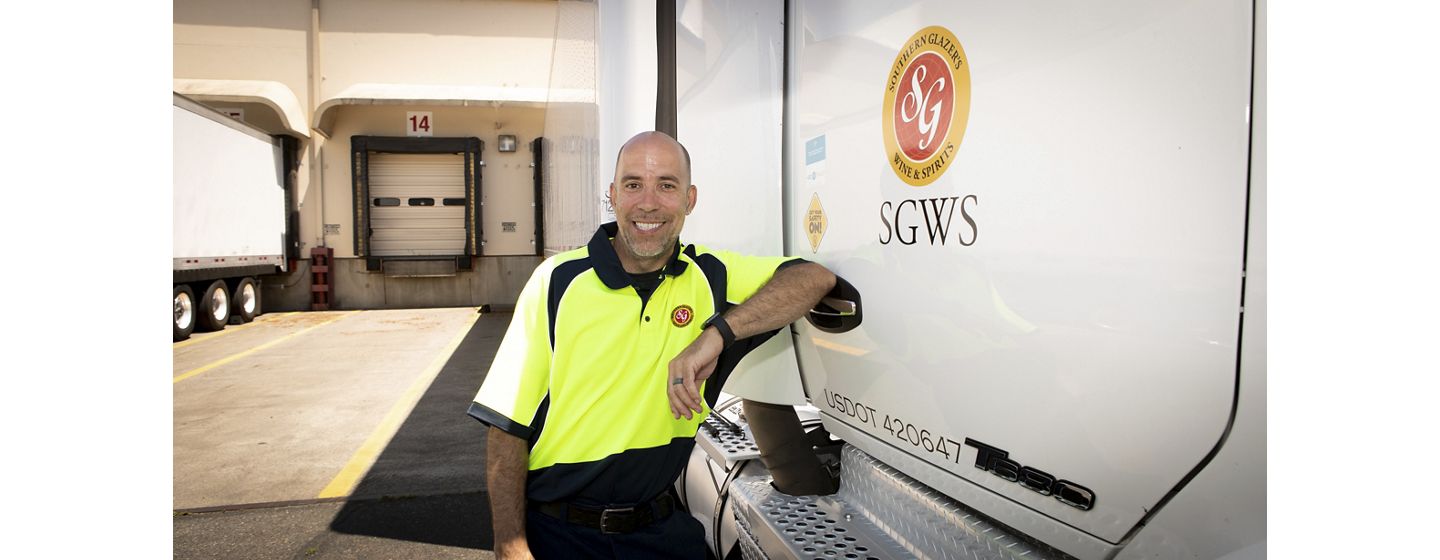 A smiling man in a neon yellow and black uniform leans on a Southern Glazer's Wine & Spirits delivery truck, exuding a friendly, professional demeanor.