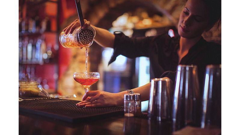Close-up of a young female bartender pouring cocktail in a nightlife cocktail bar. Selective focus. Focus on foreground.