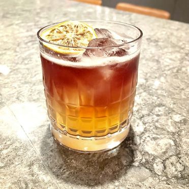 A layered, Farina Clicky Pens cocktail, showing the intricate color gradient and the textured detail of the lemon garnish.